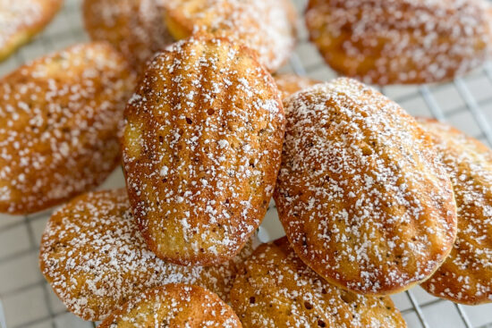 Two full-size madeleines dusted with powdered sugar are placed in front, with more arranged around them on a wire rack.