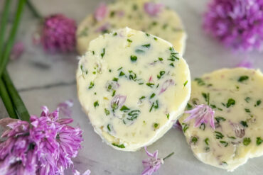 In the main view, one slice of chive blossom butter is clearly visible, with two more slices in the background and several chive blossoms scattered around. The slices are placed on a white and grey marble surface.