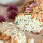 Garlic Chive Cheese Log covered with chopped walnuts with two slices in front of the log, placed on parchment paper and surrounded by a few chive blossoms and fresh greens. In the top left corner, a white bowl filled with chive blossoms sits beside a white plate holding a few crackers. On the right side, next to the cheese slices, one cracker is topped with the cheese spread.