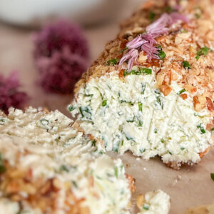 Garlic Chive Cheese Log covered with chopped walnuts with two slices in front of the log, placed on parchment paper and surrounded by a few chive blossoms and fresh greens. In the top left corner, a white bowl filled with chive blossoms sits beside a white plate holding a few crackers. On the right side, next to the cheese slices, one cracker is topped with the cheese spread.
