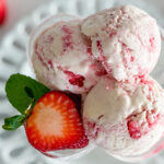 Three scoops of strawberry ice cream and a halved fresh strawberry are arranged in a glass dessert bowl, viewed from the top. The bowl sits on a white porcelain plate with a decorative trim around the edges. A few strawberries are scattered casually beside the plate.
