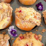 A dark grey muffin pan partially filled with freshly baked rhubarb muffins topped with cinnamon streusel. A few muffins are slightly tilted or lifted, resting at different angles in the pan. On the top right corner, one empty muffin cup is filled with chopped red rhubarb, adding a pop of color. Scattered around the muffins are a few delicate purple pansies.