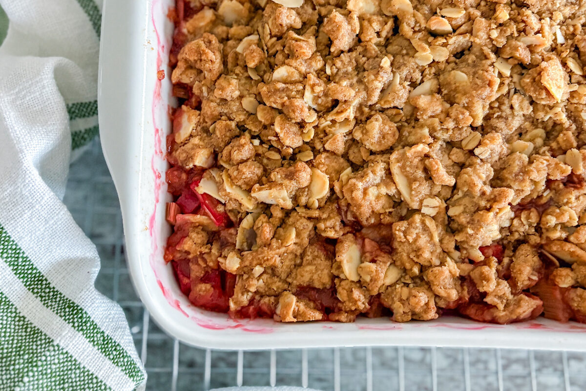 Baked rhubarb crisp with a crunchy topping of oats, almonds, and cinnamon in a white square baking dish, cooling on a wire rack. A white kitchen towel with green stripes rests beside it, all set on a grey countertop.
