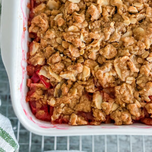 Baked rhubarb crisp with a crunchy topping of oats, almonds, and cinnamon in a white square baking dish, cooling on a wire rack. A white kitchen towel with green stripes rests beside it, all set on a grey countertop.