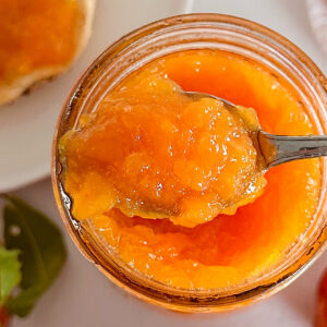 A spoonful of peach ginger jam hovers over a glass canning jar, showcasing the jam. Surrounding the jar are fresh whole peaches and a white plate with half a bagel generously spread with the golden jam. The entire scene is set on a bright white surface.