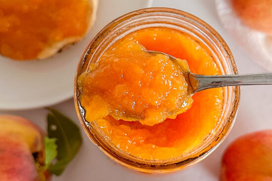 A spoonful of peach ginger jam hovers over a glass canning jar, showcasing the jam. Surrounding the jar are fresh whole peaches and a white plate with half a bagel generously spread with the golden jam. The entire scene is set on a bright white surface.
