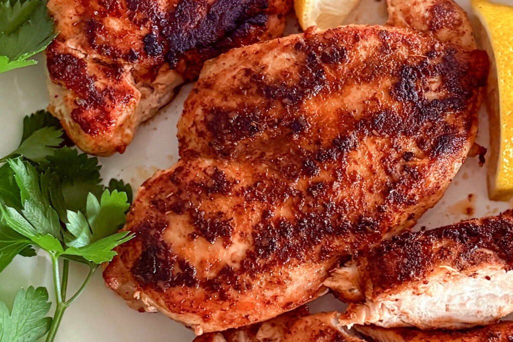 Main focus on one cooked, crisp, juicy blackened chicken breast, with a few other cooked pieces slightly visible, accompanied by a small lemon wedge and fresh parsley beside it.