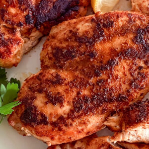 Main focus on one cooked, crisp, juicy blackened chicken breast, with a few other cooked pieces slightly visible, accompanied by a small lemon wedge and fresh parsley beside it.