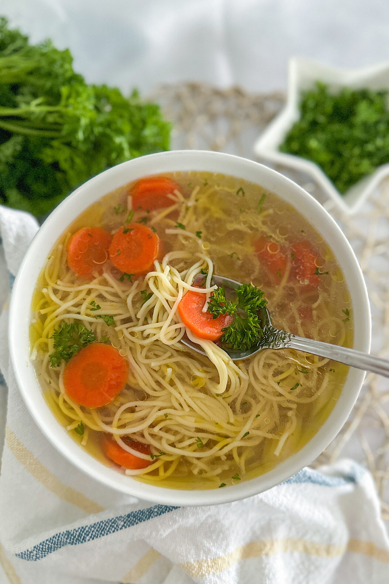 A white serving bowl filled with rosół, a clear Polish chicken soup, featuring tender noodles, sliced carrots, and a sprinkle of fresh parsley. A spoon rests over the bowl, holding noodles, a piece of carrot, and some parsley. The bowl sits on a clean white surface topped with a light gold placemat, with a white kitchen towel featuring yellow and blue stripes beside it. In the background, on the top left, is a bunch of fresh parsley, and on the opposite side, a small star-shaped dish is filled with chopped parsley.