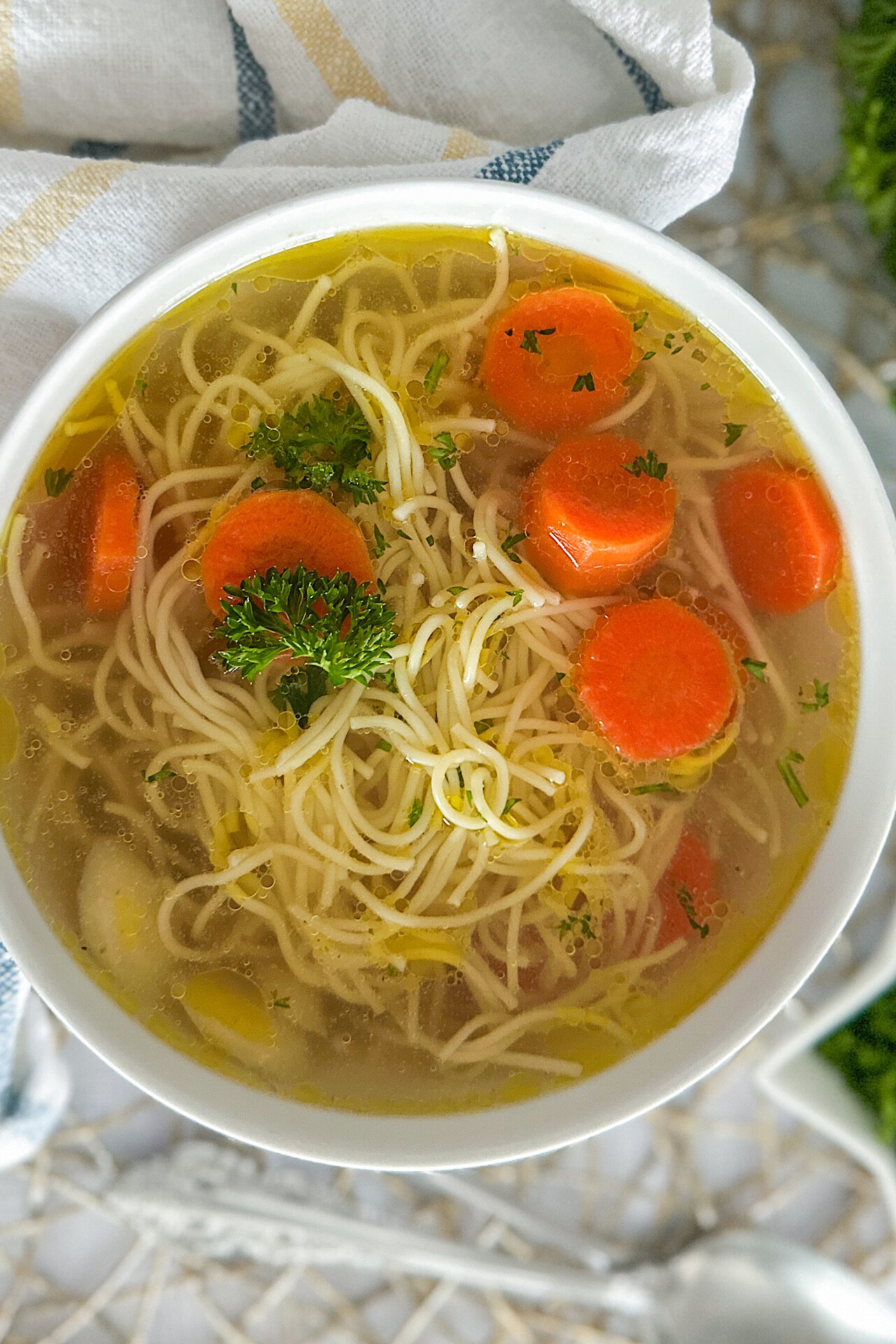A white serving bowl filled with rosół, a clear Polish chicken soup, featuring tender noodles, sliced carrots, and a sprinkle of fresh parsley as garnish. The bowl is set on a clean white surface, beside is place white kitchen towel with yellow and blue stripes. 