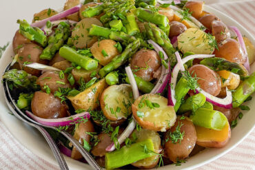Potato Asparagus Salad with red onion is tossed in lemon Dijon dressing and topped with fresh chopped dill and chives. It is served in a large round white ceramic dish with two serving spoons inside. A fresh bunch of dill rests beside the dish, which sits on a white kitchen towel with light pink stripes, all arranged on a white surface.