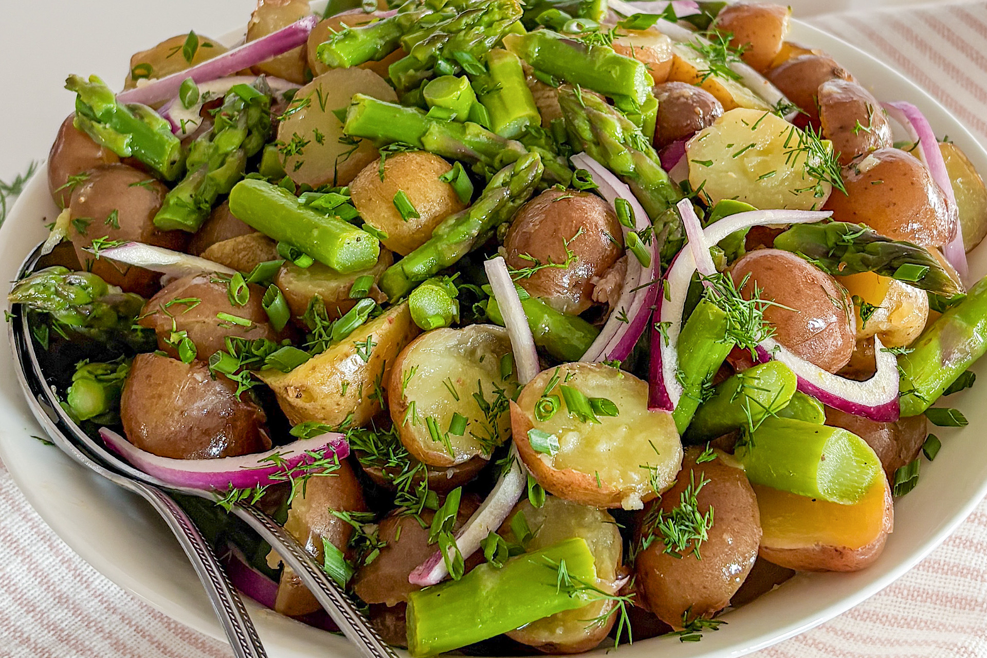 Potato Asparagus Salad with red onion is tossed in lemon Dijon dressing and topped with fresh chopped dill and chives. It is served in a large round white ceramic dish with two serving spoons inside. A fresh bunch of dill rests beside the dish, which sits on a white kitchen towel with light pink stripes, all arranged on a white surface.