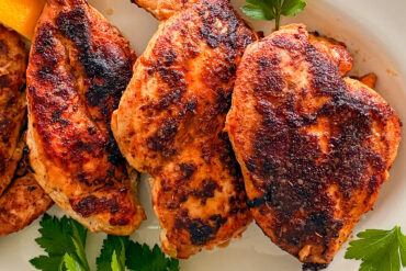 Four cooked blackened chicken breasts are arranged on a white porcelain oval serving platter, garnished with fresh parsley and accompanied by lemon pieces placed in the left corner of the picture.