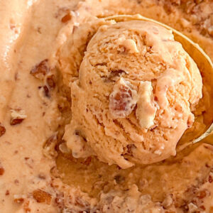 The image shows pumpkin pie ice cream topped with chopped walnuts in a light-colored loaf pan, with a gold spoonful of ice cream resting on top. The pan is placed on a white surface, beside a waffle cone and a small orange crystal pumpkin-shaped decoration.