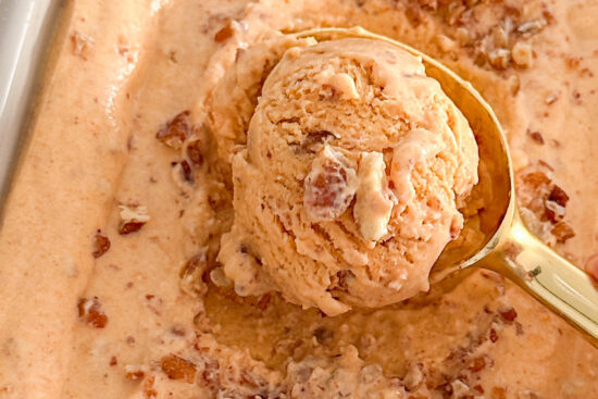 The image shows pumpkin pie ice cream topped with chopped walnuts in a light-colored loaf pan, with a gold spoonful of ice cream resting on top. The pan is placed on a white surface, beside a waffle cone and a small orange crystal pumpkin-shaped decoration.