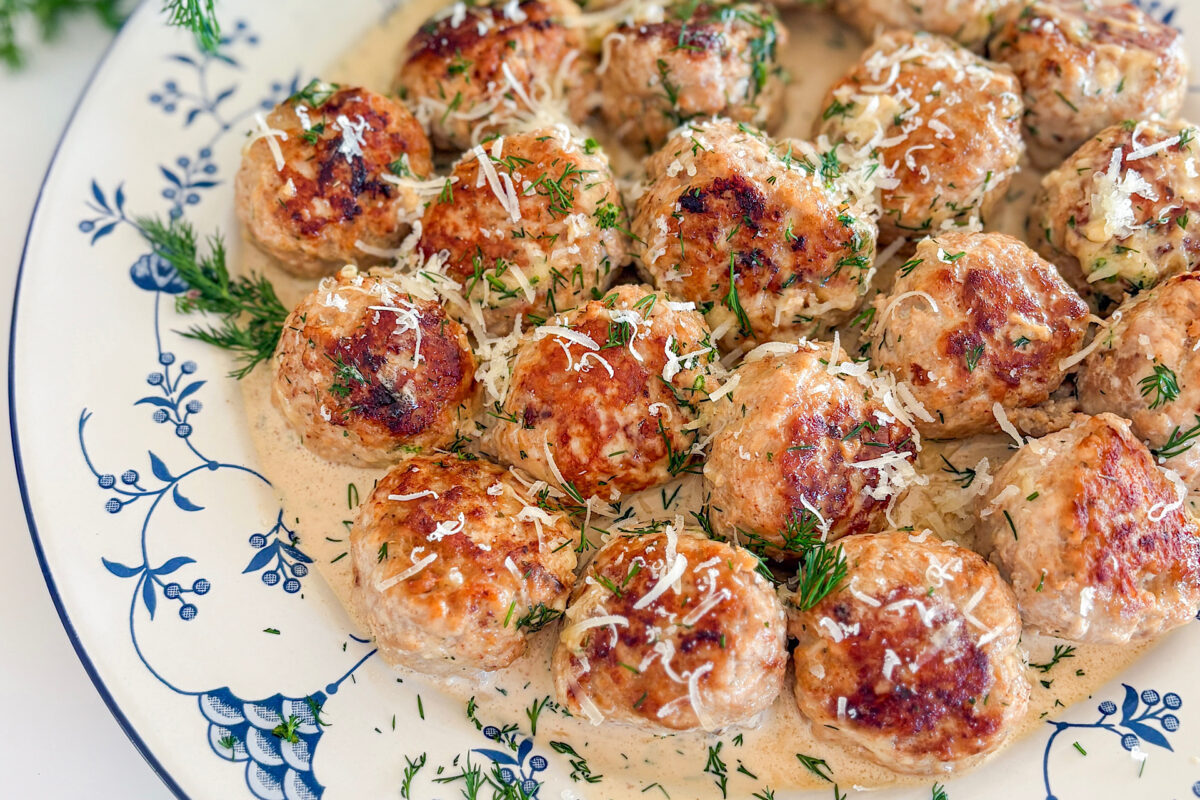 Golden brown turkey meatballs in creamy dill sauce served on a white plate with a subtle blue design. The meatballs are generously topped with freshly grated Parmesan cheese and a sprinkle of chopped fresh dil.
