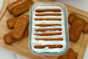 A rectangular glass container filled with plain Greek yogurt has eight Biscoff cookies inserted vertically into the yogurt. The container sits on a wooden cutting board, with additional Biscoff cookies placed on both sides of the dish.