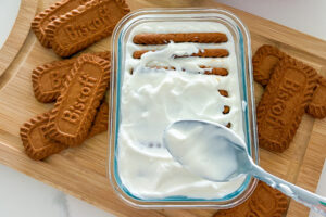 A rectangular glass container filled with plain Greek yogurt, with some Biscoff cookies visible and others fully covered by the yogurt. The container sits on a wooden cutting board, with additional Biscoff cookies placed on both sides of the dish.