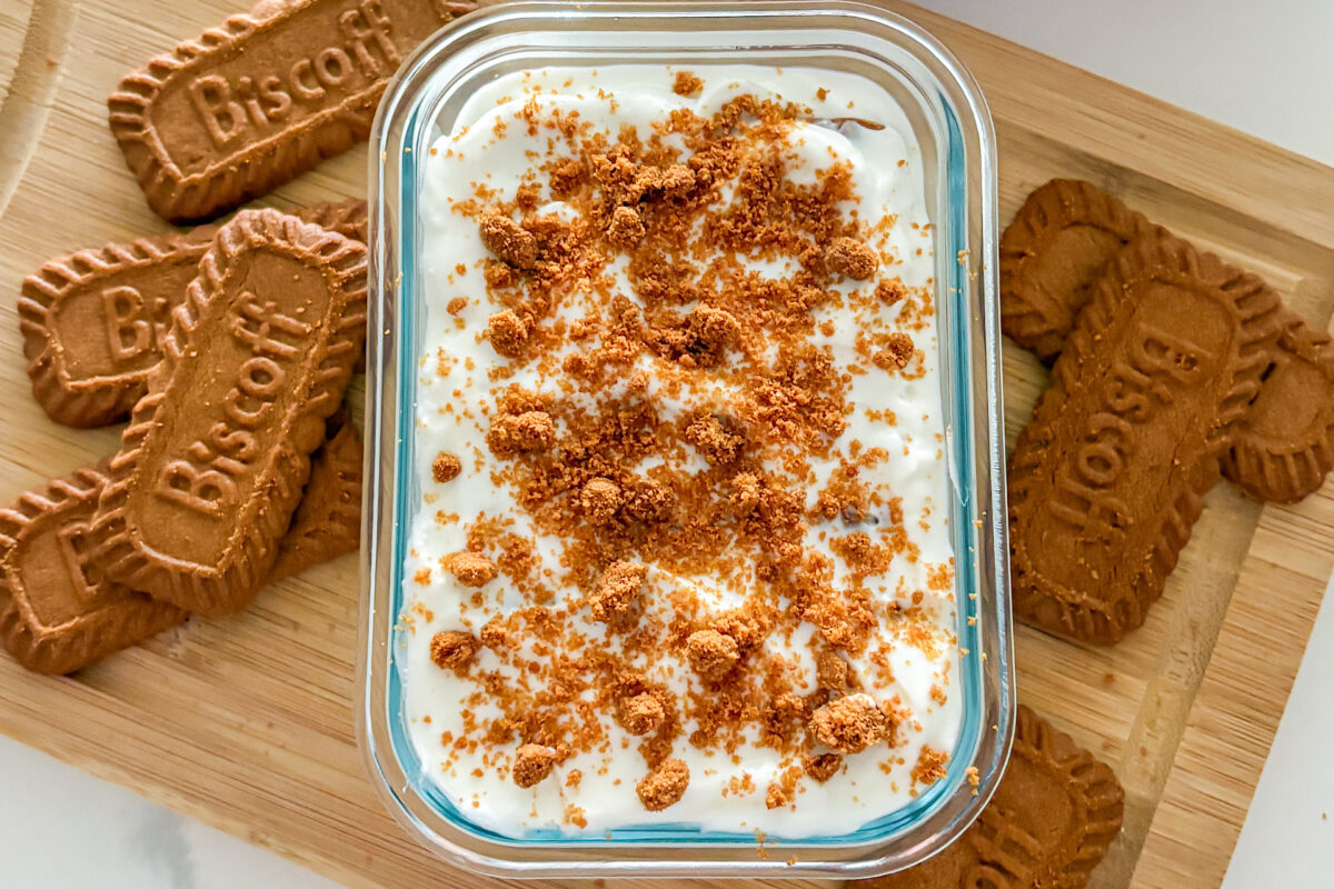 A rectangular glass container filled with plain Greek yogurt and Biscoff cookies, covered with yogurt and topped with crushed cookie crumbs. The container sits on a wooden cutting board, with additional Biscoff cookies placed on both sides of the dish, all displayed on a white kitchen counter.