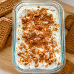 A rectangular glass container filled with plain Greek yogurt and Biscoff cookies, covered with yogurt and topped with crushed cookie crumbs. The container sits on a wooden cutting board, with additional Biscoff cookies placed on both sides of the dish, all displayed on a white kitchen counter.