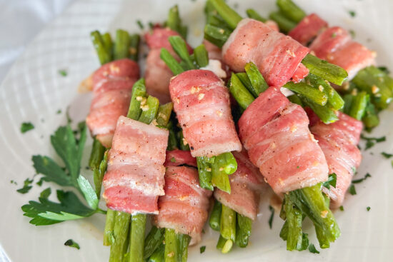 A bunch of easy to make, savory side dish of baked bacon wrapped spicy beans wrapped in bacon bundles, topped with chopped parsley, and served on a white oval porcelain serving plate set on a white surface.