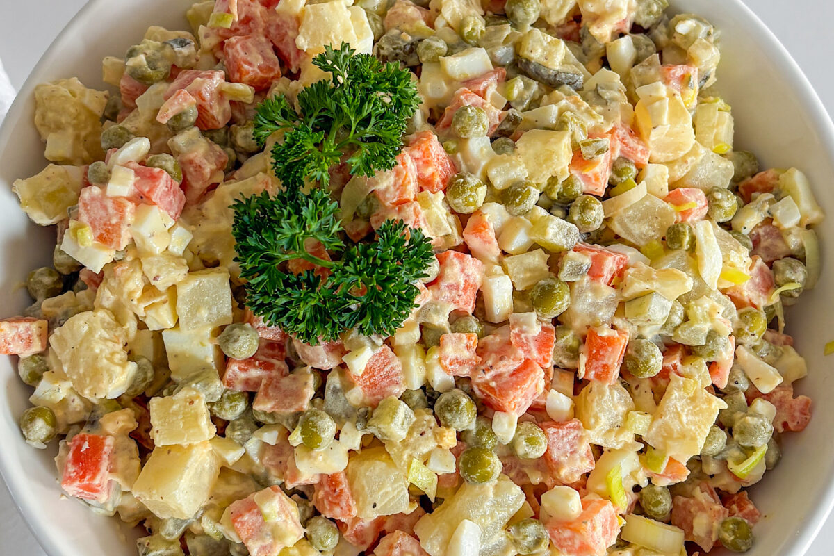 Creamy, mayo-based Polish vegetable salad, fully mixed and served in a white porcelain bowl, garnished on top with a sprig of fresh parsley. the serving bowl is placed on white surface.