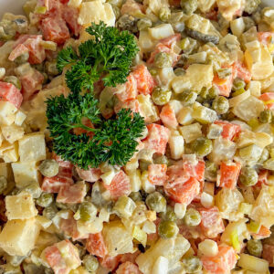 Creamy, mayo-based Polish vegetable salad, fully mixed and served in a white porcelain bowl, garnished on top with a sprig of fresh parsley. the serving bowl is placed on white surface.