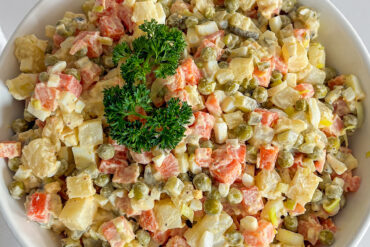 Creamy, mayo-based Polish vegetable salad, fully mixed and served in a white porcelain bowl, garnished on top with a sprig of fresh parsley. the serving bowl is placed on white surface.