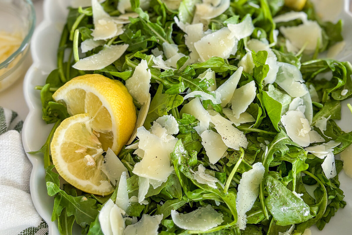 Arugula salad is mixed and placed on a white porcelain serving plate. The top is sprinkled with shaved Parmesan cheese flakes and mixed with lemon honey Vinaigrette. The top of the salad is sprinkled with fresh ground Black pepper. On the left side, near the edge of the plate, there are two slices of fresh lemon, placed over the salad. In the top left corner, a small glass bowl filled with pine nuts is partially visible. A white kitchen towel with light green stripes is placed next to the plate.