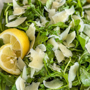 Arugula salad is mixed and placed on a white porcelain serving plate. The top is sprinkled with shaved Parmesan cheese flakes and mixed with lemon honey Vinaigrette. The top of the salad is sprinkled with fresh ground Black pepper. On the left side, near the edge of the plate, there are two slices of fresh lemon, placed over the salad. In the top left corner, a small glass bowl filled with pine nuts is partially visible. A white kitchen towel with light green stripes is placed next to the plate.