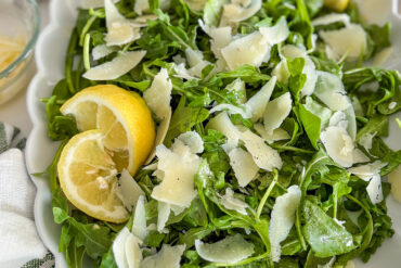 Arugula salad is mixed and placed on a white porcelain serving plate. The top is sprinkled with shaved Parmesan cheese flakes and mixed with lemon honey Vinaigrette. The top of the salad is sprinkled with fresh ground Black pepper. On the left side, near the edge of the plate, there are two slices of fresh lemon, placed over the salad. In the top left corner, a small glass bowl filled with pine nuts is partially visible. A white kitchen towel with light green stripes is placed next to the plate.