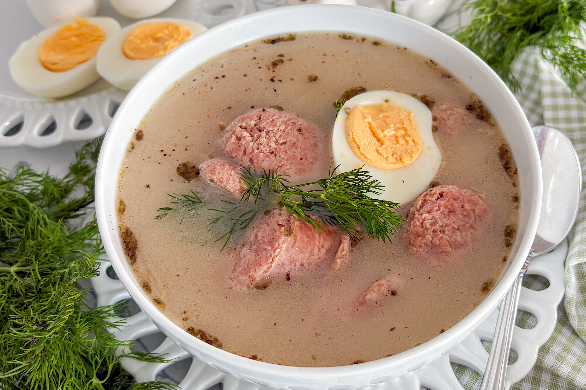 A white porcelain serving bowl filled with Polish sour soup Żurek, containing pieces of white sausage and half a boiled egg, and garnished with fresh dill, sits on a matching porcelain plate with lace-designed edges. A stainless steel spoon rests beside the bowl. In the top left corner, a small white plate with the same lace edge is placed slightly below. More fresh dill is arranged in the top right corner. To the bottom right of the bowl, a folded fabric napkin in white with a light green pattern sits beside the plate. The entire setting rests on a white tablecloth with delicate light green floral designs.