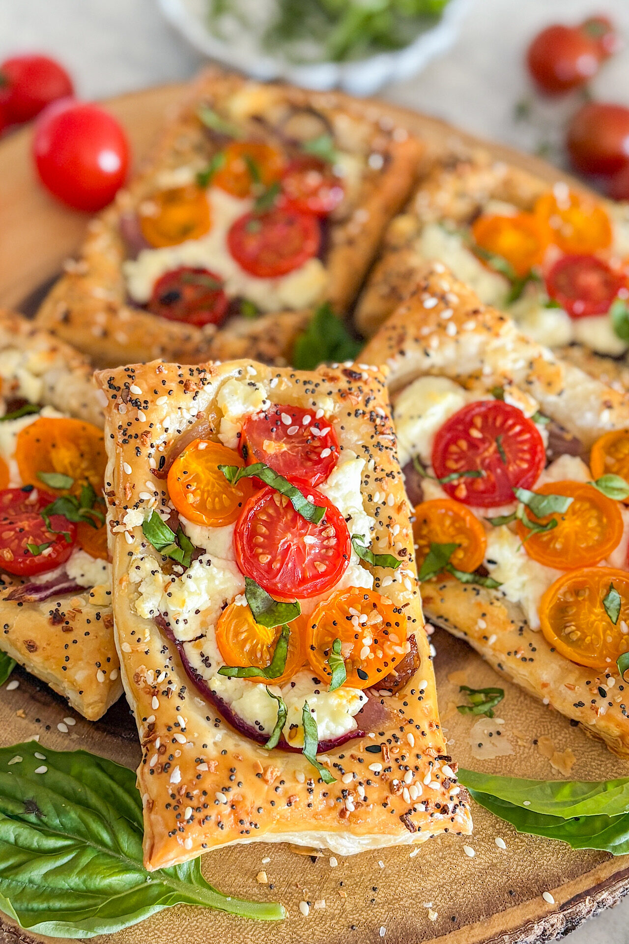 In the main view, one baked puff pastry tart is visible, topped with caramelized red onion, creamy melted goat cheese, and fresh basil. The crunchy puff pastry is sprinkled with everything bagel seasoning. Beside it, four more tart pieces are placed, some closer and more in focus, while others are slightly blurred in the background. In the front at the bottom of the picture, two fresh basil leaves are placed. In the background, small cherry tomatoes are positioned in the top left and top right corners. In the top middle of the image, a small serving bowl with freshly chopped basil is partially visible. The tarts are arranged on a round brown cutting board sprinkled with seasoning. Everything is placed on a white surface.