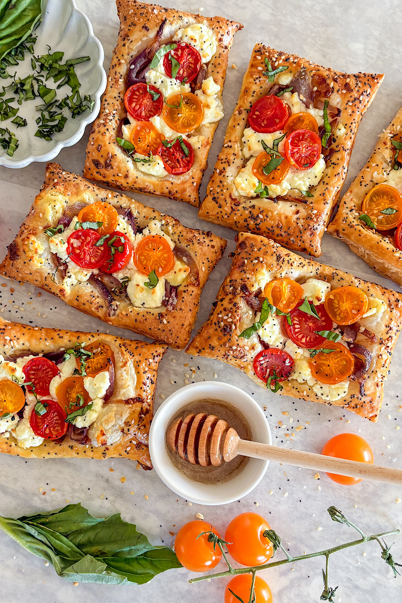 Five puff pastry cherry tomato goat cheese tarts are fully visible, with one more partially visible in the top right corner. In the lower part of the picture is a branch with yellow cherry tomatoes attached, along with fresh basil leaves and a small white serving bowl filled with honey and a honey spoon. In the top left corner, a light grey small plate with chopped basil is visible. The whole display is arranged on a white surface.