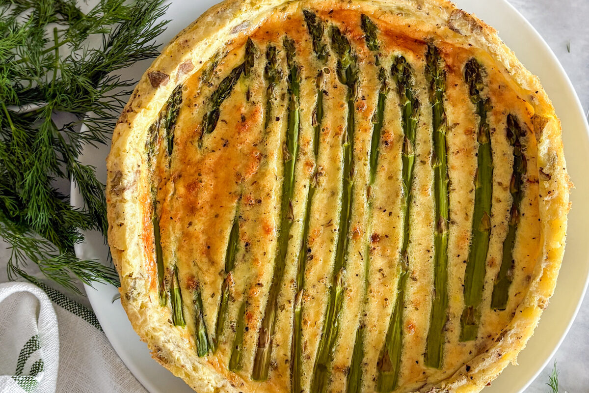 A baked asparagus quiche is served on a white plate. At the bottom of the image, beside the plate, is a small glass bowl filled with shredded Parmesan cheese. On the opposite side, a folded white kitchen towel with green stripes adds a soft accent. Sprigs of fresh dill are scattered around the plate. The composition is set on a light grey surface.