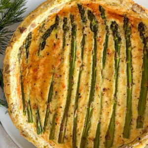 A baked asparagus quiche is served on a white plate. At the bottom of the image, beside the plate, is a small glass bowl filled with shredded Parmesan cheese. On the opposite side, a folded white kitchen towel with green stripes adds a soft accent. Sprigs of fresh dill are scattered around the plate. The composition is set on a light grey surface.