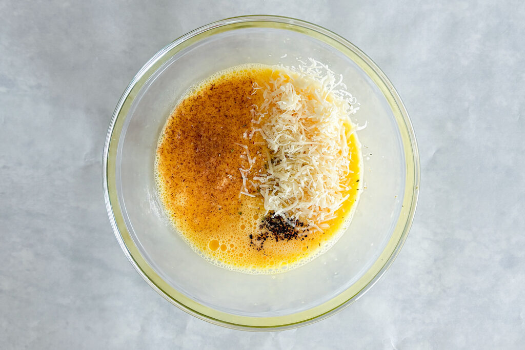 The filling for the asparagus quiche is in a glass bowl. The mixture contains eggs, milk, Parmesan cheese, nutmeg, oregano, salt, and pepper. The glass bowl is placed on a light grey surface.